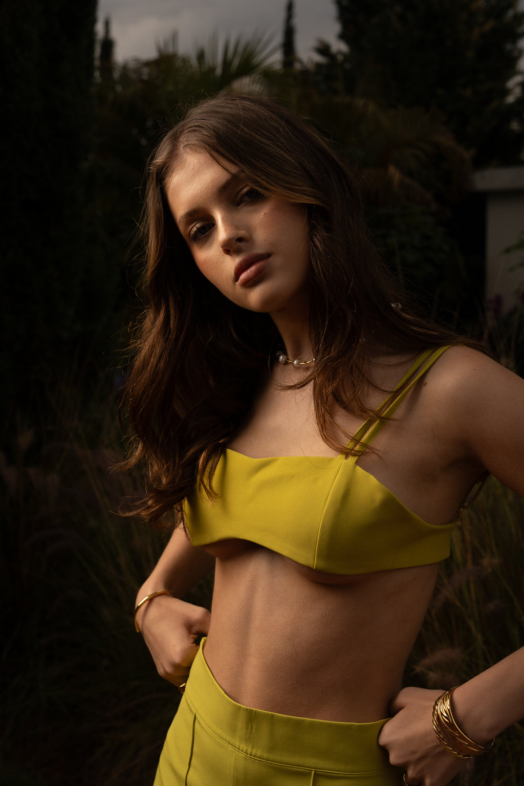 Model wearing Conventus IV Mini Top and pants in green structured mini cropped silhouette paired with gold jewelry standing in the garden of a luxury italian villa.