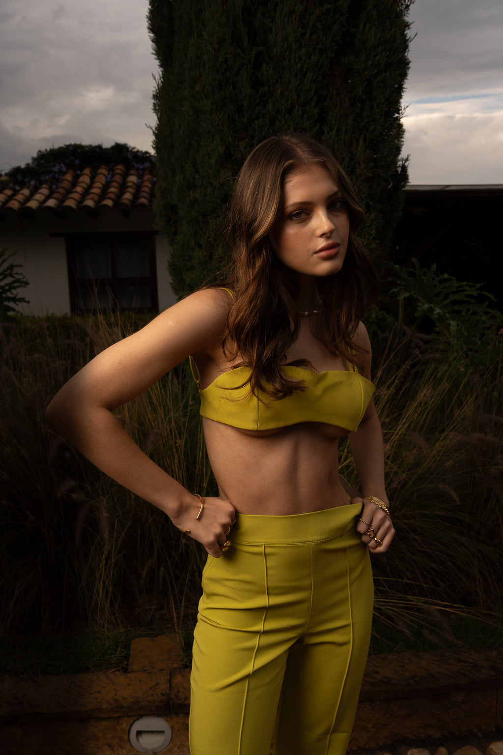 Full‑length of a model wearing Conventus IV Pants and mini top featuring swirling green pattern, high-waisted and wide‑leg cut. Standing in the garden of a luxury italian villa.