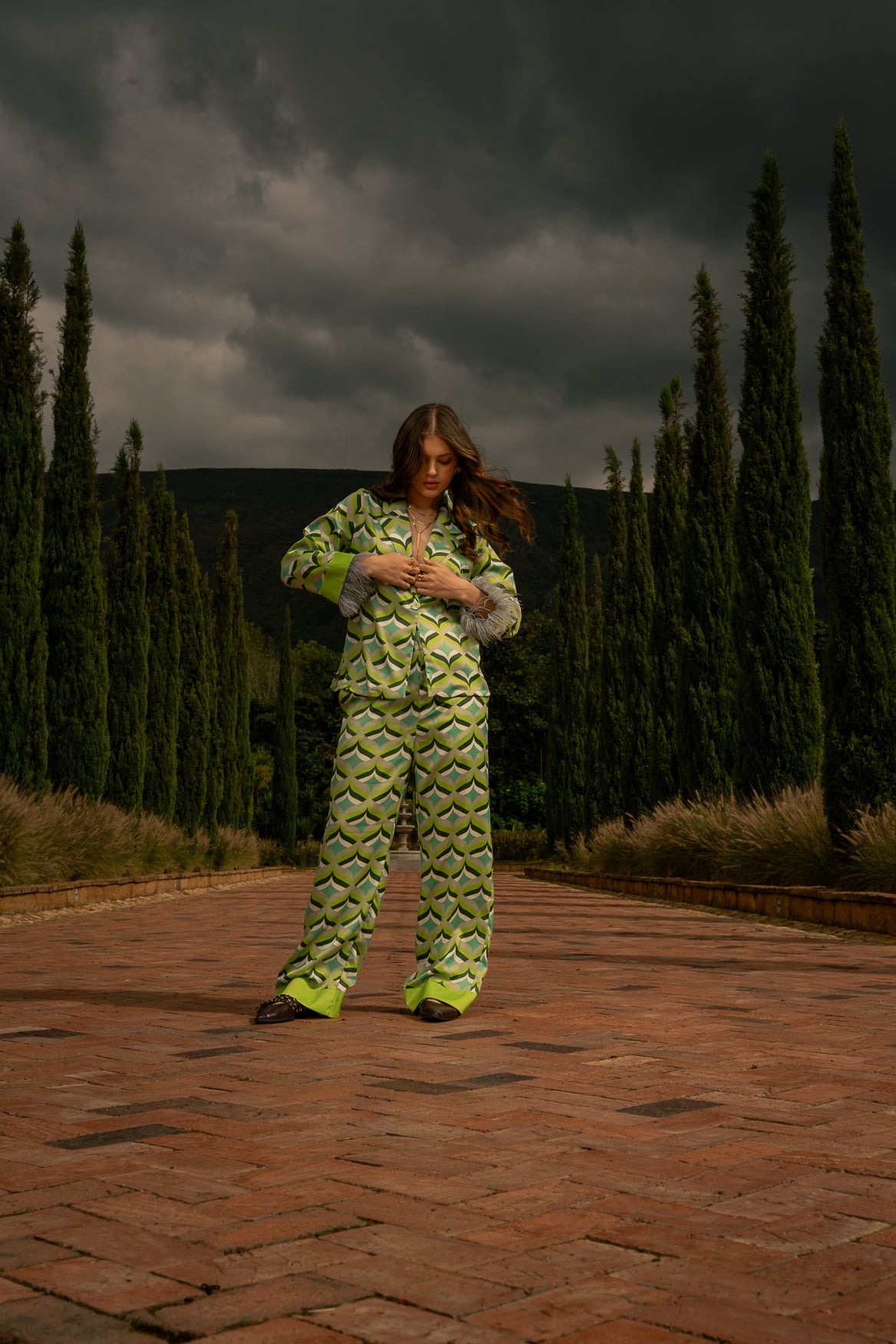Model on a cobblestone path wearing Vita I Pants and matching Vita I blouse with feathered cuffs framed by cypress trees. Old money aesthetic.