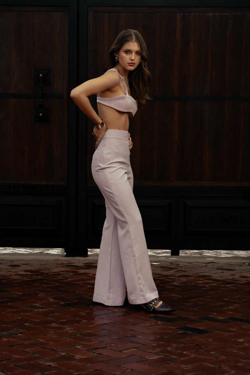 Woman wearing blush‑toned Fuga III Pants, high‑waisted and tailored, standing outdoors in a luxury italian villa entrance.