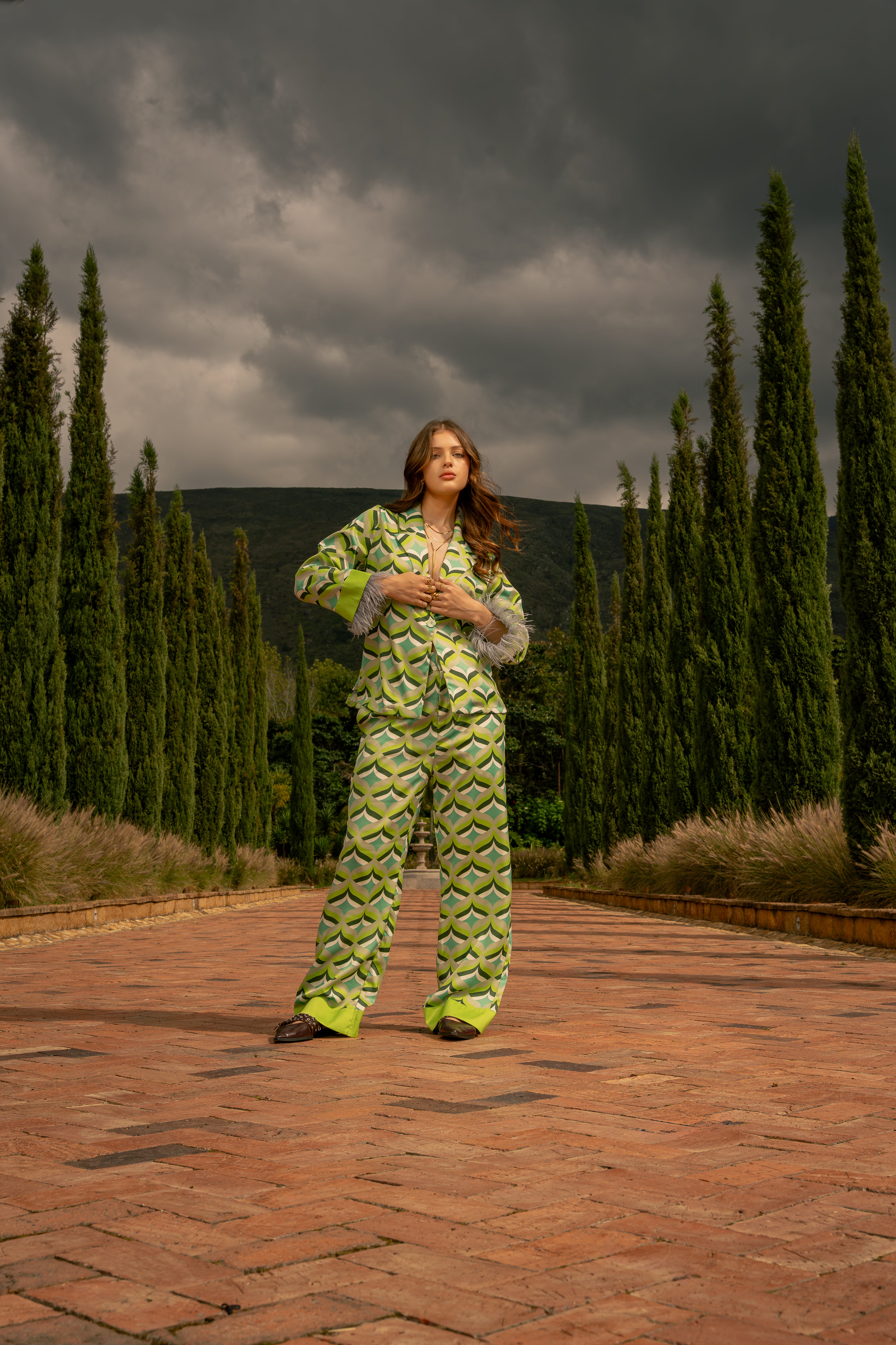 Model on a cobblestone path wearing Vita I Pants and matching Vita I blouse with feathered cuffs framed by cypress trees. Old money aesthetic.