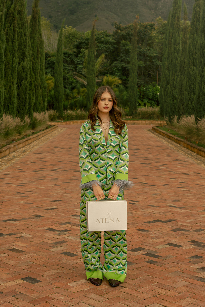 Model wearing Vita I Blouse and Vita I Pants in green geometric print, holding an ATENA box  in a luxury italian villa garden path surrounded of cypress trees. Old money aesthetic 
